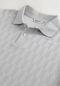 Polo gris clair avec un motif géométrique texturé subtil et deux boutons blancs, posé à plat montrant le col et l’étiquette au niveau du col.