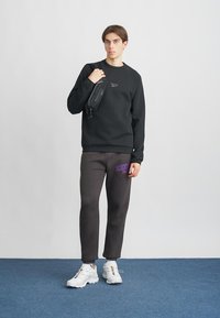 Sudadera negra de cuello redondo, pantalones de chándal grises con logo púrpura, zapatillas blancas y pequeña bolsa negra de hombro. Fondo liso y suelo azul.
