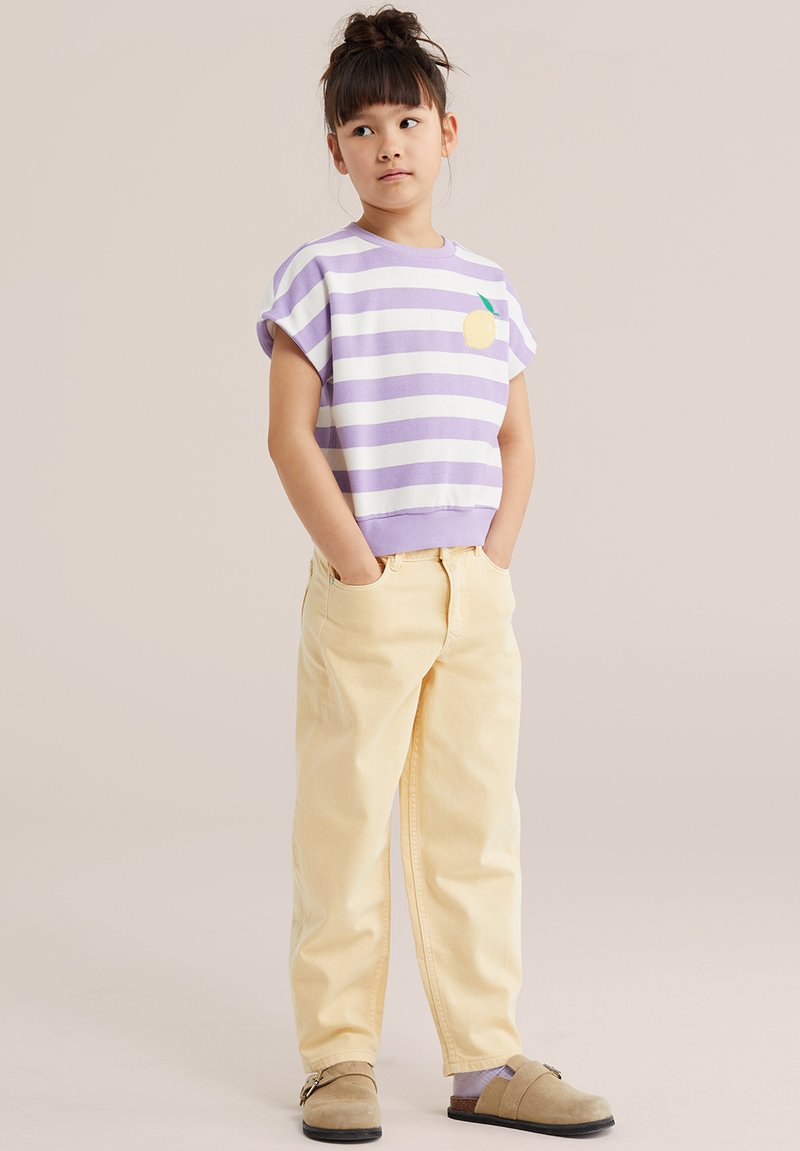 Ragazza in piedi con le mani nelle tasche, indossa una maglietta a righe lavanda e bianche con grafica di limone, pantaloni gialli e scarpe slip-on beige.