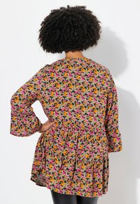 Beige jurk met bloemenpatroon en roze, oranje en groene accenten. Heeft lange mouwen en een uitlopende, gelaagde zoom.