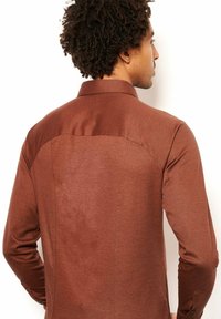 Bruine blouse met lange mouwen en een gladde textuur, voorzien van een contrasterende donkere yoke en subtiele stiksels aan de achterkant.