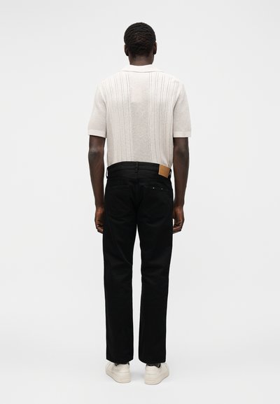 Άνδρας στέκεται με την πλάτη, φοράει κρεμ πλεκτό πόλο με κοντά μανίκια, μαύρο παντελόνι με ίσια γραμμή και λευκά αθλητικά παπούτσια σε λευκό φόντο.