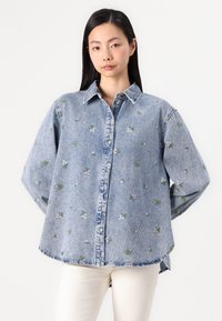 Τζιν πουκάμισο με κουμπιά και floral κεντήματα σε πράσινο και ροζ. Διαθέτει μυτερό γιακά και χαλαρή εφαρμογή, με στρογγυλή άκρη.