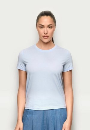 Kobieta z ciemnymi włosami związanymi w kucyk, ubrana w jasnoniebieską koszulkę z krótkim rękawem i wysoko taliowaną niebieską spódnicę, stoi na tle jednolitego białego tła.