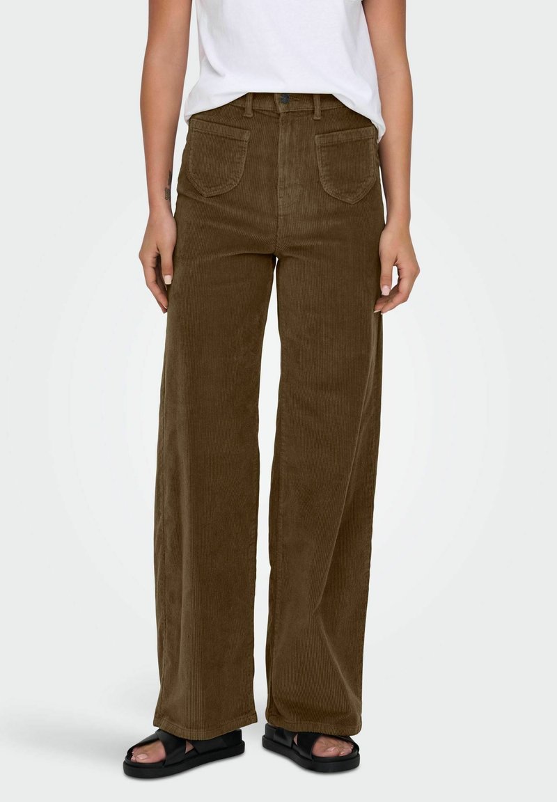 Personne portant un pantalon en velours côtelé marron à jambes larges avec des poches plaquées à l'avant, une chemise blanche rentrée, et des sandales noires à bouts ouverts, debout devant un fond uni.