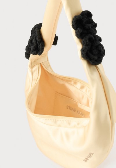 Τσάντα από κρεμ σατέν με μαύρες πλεκτές floral διακοσμήσεις στις χειρολαβές, ανοιχτό φερμουάρ στην κορυφή και το όνομα της μάρκας "STINE GOYA" εσωτερικά και εξωτερικά.