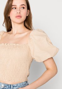 Haut beige à manches bouffantes avec un décolleté carré, un corps smocké texturé et une longueur cropped ; tissu léger et aérien avec un tombé doux.