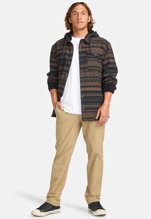 Brown patterned shirt jacket with a hood, buttoned front, and dark accents. Paired with beige pants and black sneakers. White T-shirt underneath.