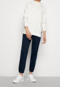 Vit, långärmad tröja med hög krage, tillsammans med marinblå smalare byxor och vita sneakers. Tyget ser mjukt och avslappnat ut.