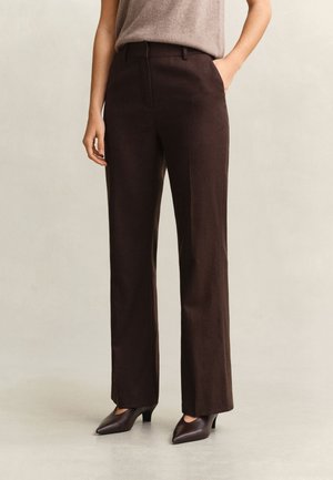 Personne portant un pantalon droit marron foncé et des talons bruns à bout pointu, debout devant un fond uni.