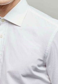 Homme portant une chemise blanche à col boutonné, visible du cou jusqu'au milieu de la poitrine sur un fond uni.