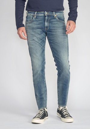 Jeans en denim bleu avec un design décoloré, dotés de cinq poches, d'une fermeture à bouton et d'une coupe slim, associés à des baskets montantes noires.