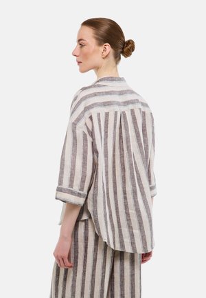 Femme aux cheveux attachés en chignon, vêtue d'une chemise ample à rayures verticales beiges et marron avec des boutons et d'un pantalon assorti, tournée vers la gauche sur un fond blanc.