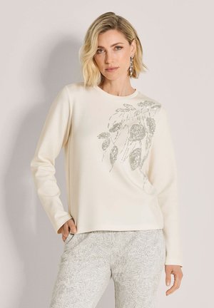Frau mit kurzen blonden Haaren, die ein cremefarbenes Langarmoberteil mit silbernen Blumenpailletten und helle gemusterte Hosen vor einem einfarbigen Hintergrund trägt.