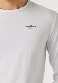 Maglietta bianca a maniche lunghe con scollo rotondo e logo "Pepe Jeans London" nero sul lato sinistro del petto, indossata da una persona.