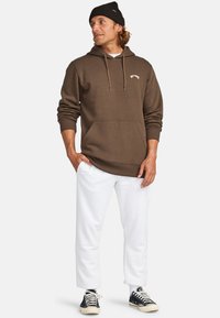 Brauner Hoodie mit Kordelzügen, Fronttasche und Logo; kombiniert mit weißen Jogginghosen und schwarzen Sneakern. Lässiges Sportoutfit.