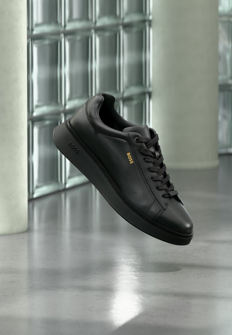 Ένα μαύρο δερμάτινο sneaker χαμηλού προφίλ με κορδόνια, χρυσό λογότυπο "BOSS" στο πλάι, εμφανίζεται να αιωρείται σε εσωτερικό χώρο με φόντο γυάλινους τούβλους και σκυρόδεμα.