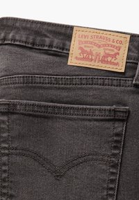 Jeans de mezclilla gris con un parche de cuero marrón claro que tiene un texto rojo y un gráfico de caballos. La costura es de color marrón oscuro con su diseño característico en el bolsillo trasero.