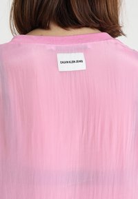 Różowa półprzezroczysta bluzka z okrągłym dekoltem i metką "CALVIN KLEIN JEANS" z tyłu, cechująca się gładką teksturą i delikatnym połyskiem.