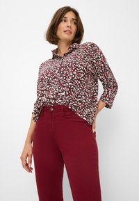 Blouse rouge foncé avec un motif floral blanc et rose clair, dotée d'un col et de manches trois-quarts, accompagnée de pantalons rouges foncés.