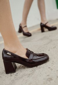 Mocasines de Charol burdeos con tacón ancho, que cuentan con una tira horizontal en la parte superior y un pequeño acento metálico.