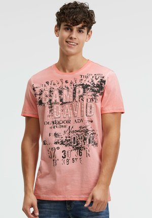 Junger Mann mit lockigem Haar, der ein verblasstes korallenrotes T-Shirt mit Grafik und blaue Jeans trägt, lächelnd vor neutralem Hintergrund.