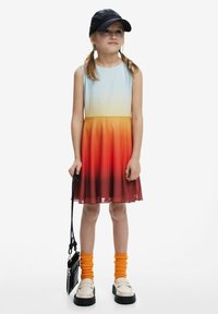 Ein ärmelloses Kleid mit Farbverlauf von Hellblau zu Orange, mit einem strukturierten Saum, kombiniert mit orangefarbenen Socken und schwarzen Schuhen. Schwarze Mütze und Tasche.