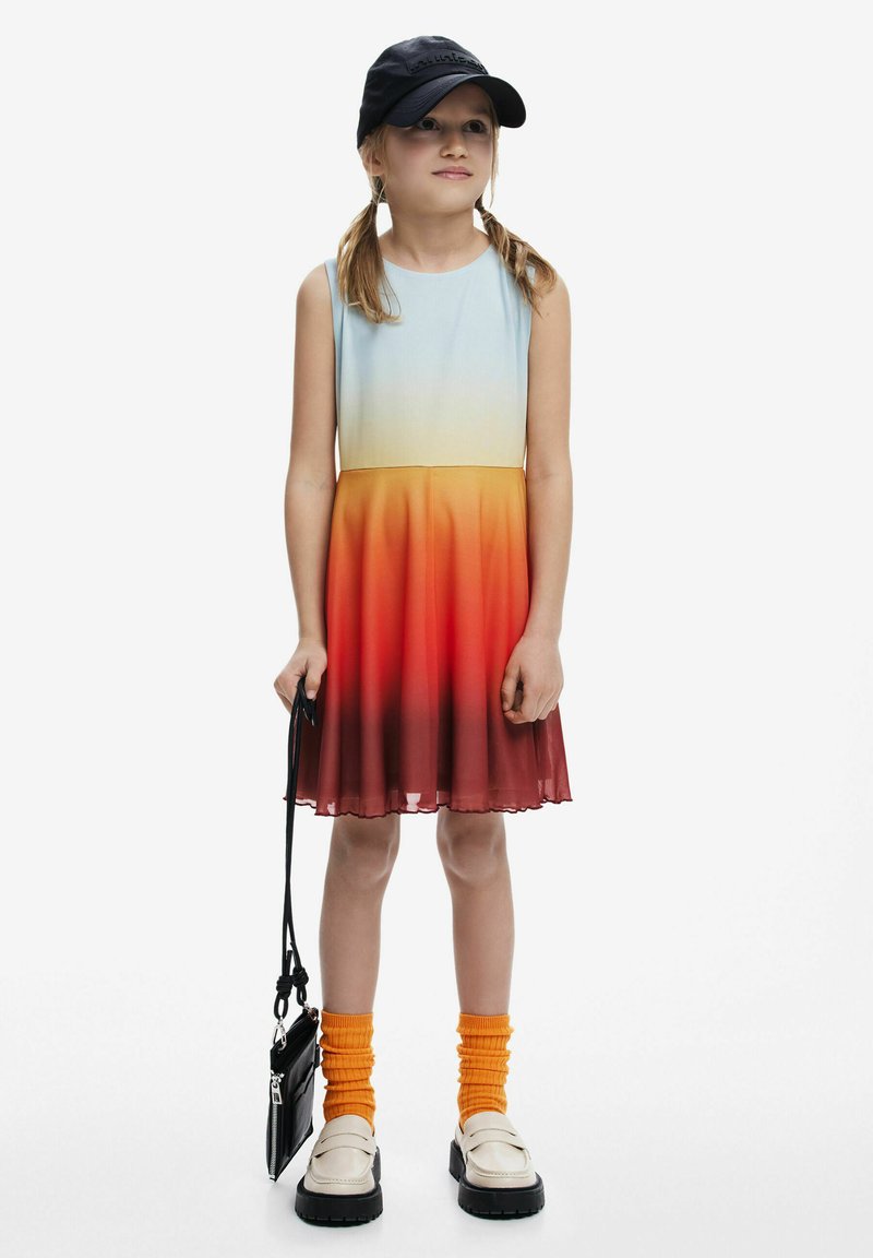 Ein ärmelloses Kleid mit Farbverlauf von Hellblau zu Orange, mit einem strukturierten Saum, kombiniert mit orangefarbenen Socken und schwarzen Schuhen. Schwarze Mütze und Tasche.
