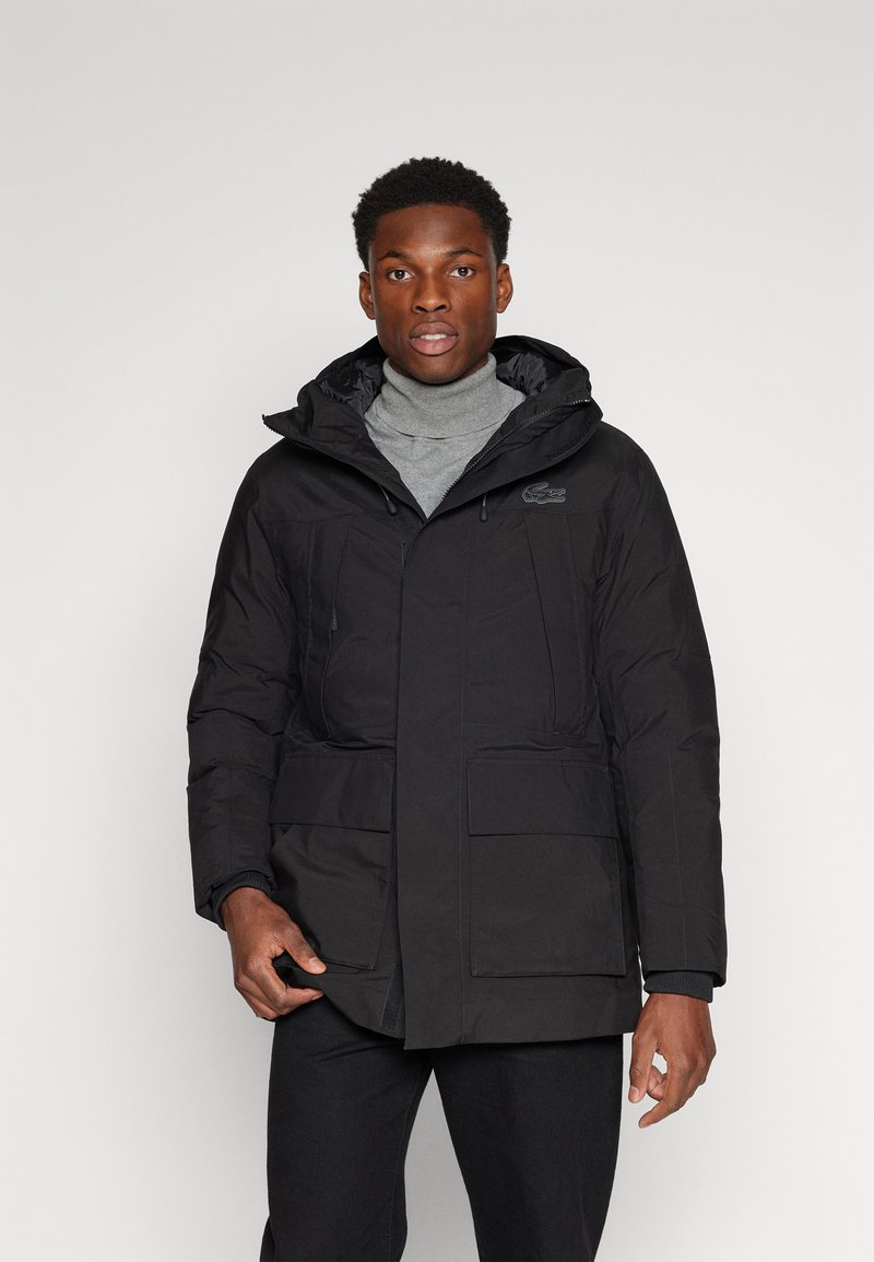 Chaqueta acolchada negra con capucha, que cuenta con dos bolsillos frontales, un cierre de cremallera y un pequeño logo en el pecho. Llevada sobre un suéter gris de cuello alto.