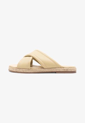 Tan leather slide sandal with crisscross straps, featuring a natural fiber sole and a flat, rounded shape. Smooth texture with minimal detailing.