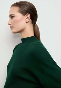 Dunkelgrüner, hochschlüssiger Pullover mit lockerer Passform. Der Stoff wirkt glatt, hat lange Ärmel und ein minimalistisches Design. Die Haare sind zu einem glatten Pferdeschwanz gebunden.
