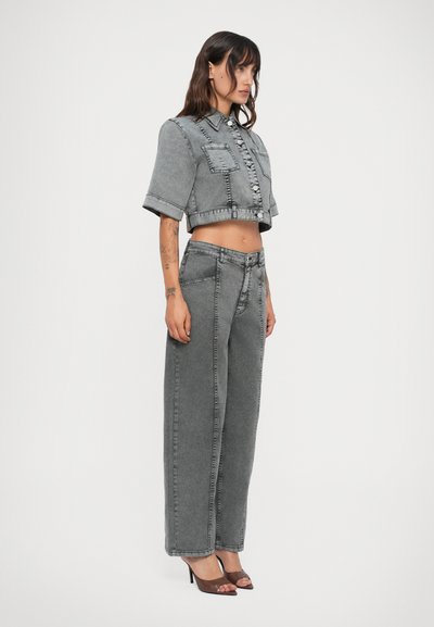 Kvinne iført grå kort jakke og høy midje bredbente jeans, brune sandalhæler, står foran en nøytral studio bakgrunn.