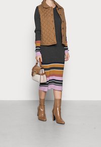 Chaleco acolchado marrón sobre un vestido de punto gris con rayas multicolores. Botas marrones texturizadas y un bolso pequeño con acentos.