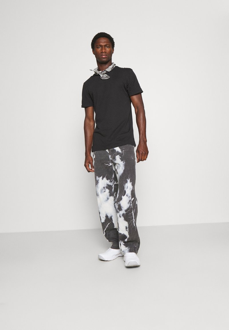 Svart t-skjorte, grå og hvite tie-dye jeans, og hvite sneakers; modell står med avslappede hender langs siden, enkel bakgrunn.