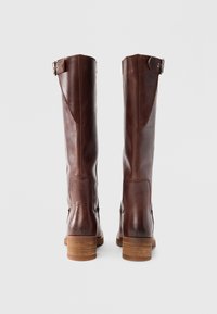 Καφέ δερμάτινες μπότες έως το γόνατο με λεία υφή, δύο διακοσμητικές λουρίδες στο πάνω μέρος και χοντρό ξύλινο τακούνι.