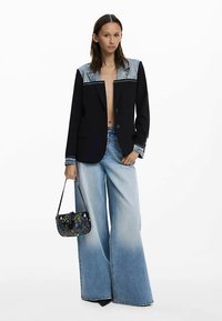 Czarna marynarka z jeansowymi dodatkami, zestawiona z szerokimi niebieskimi dżinsami. Trzyma małą torebkę w wzory. Białe tło.