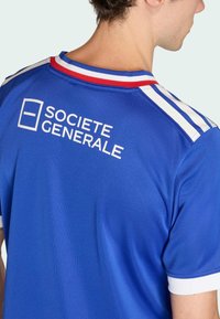 Modrý sportovní dres s kulatým červeno-bílým límcem, bílými pruhy na ramenou a nápisem „SOCIETE GENERALE“ bílou barvou na zádech.