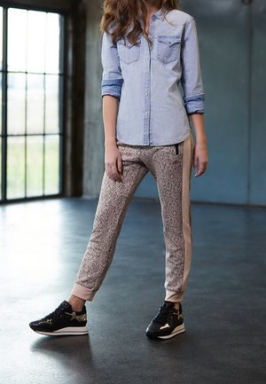 Vrouw die binnen staat, gekleed in een licht denim shirt, sportieve broek met zijstrepen en zwarte sneakers met plateauzool.