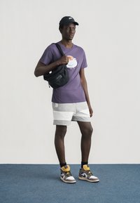 Νέος άνδρας στέκεται σε δάπεδο με χαλί, φοράει μοβ μπλουζάκι με γραφική απεικόνιση φλιτζανιού καφέ, γκρι σορτς, μαύρο καπέλο, τσάντα χιαστί και ψηλά αθλητικά παπούτσια.