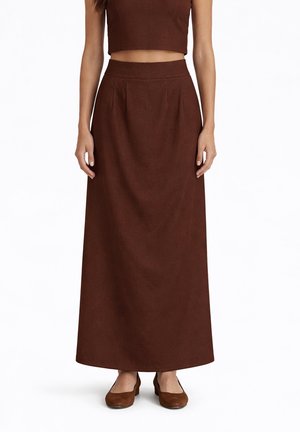 Donna che indossa una gonna marrone a vita alta, lunga fino alla caviglia, con una cintura aderente e scarpe basse marroni abbinate, in piedi davanti a uno sfondo bianco.