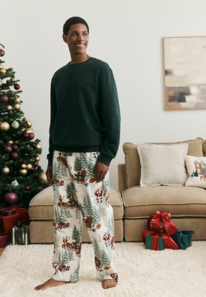 Man smiling indoors wearing a dark green sweatshirt and Christmas-themed pajama pants near a decorated tree and wrapped gifts.