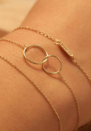Bracelet en chaîne dorée avec deux cercles entrelacés et une barre ornée de pierres claires, posé sur une peau bronzée pour créer un contraste.