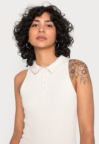 Top de polo sem mangas, em creme, com um padrão nervurado, colarinho e botão. Apresenta um tatuagem floral no ombro.