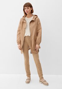 Lekka kurtka z kapturem w kolorze beżowym, z elastycznymi mankietami, przednimi kieszeniami i zamkiem błyskawicznym. W zestawie ze dopasowanymi spodniami i butami wsuwanymi.