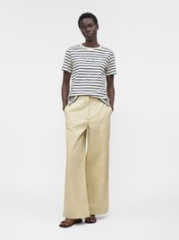 Femme portant un t-shirt rayé "Weekend", un pantalon large beige et des sandales marron, debout avec les mains dans les poches devant un fond uni.