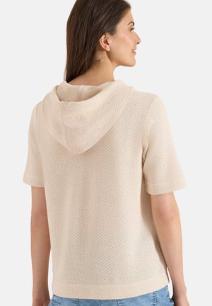 Kvinde iført en beige strikket kortærmet hoodie, vist bagfra, med langt brunt hår og lyseblå jeans.