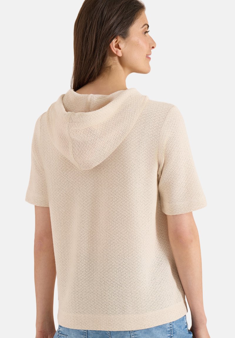 Femme portant un hoodie beige en maille à manches courtes, vue de dos, avec de longs cheveux bruns et un jean bleu clair.