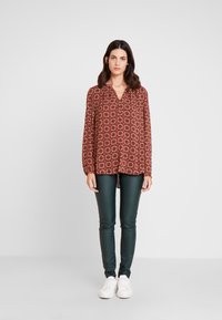 Blusa vermelha com padrão e mangas longas, decote em v e parte traseira ligeiramente mais longa. Combinada com leggings de couro verde-escuro e ténis brancos.