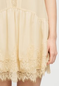 Μπεζ φόρεμα με στρώσεις λεπτομερειών δαντέλας στο τελείωμα, υλικό που μαζεύεται πάνω από τη δαντέλα και ένα χέρι που ακουμπάει δίπλα στη φούστα.