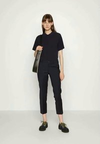 Chemise noire à manches courtes, pantalon noir ajusté et chaussures noires massives avec des lacets jaunes. Sac à bandoulière noir avec une longue sangle. Fond simple.